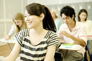 study-in-canada Five young students sitting at desks in a classroom or lecture hall, with a smiling female Asian student in the foreground